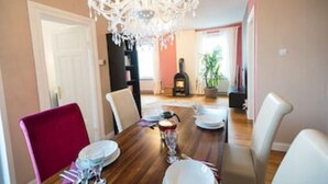 Un coin repas avec une table en bois dressée pour quatre personnes, entourée de chaises. Un lustre est suspendu au plafond, et une cheminée est visible au fond.