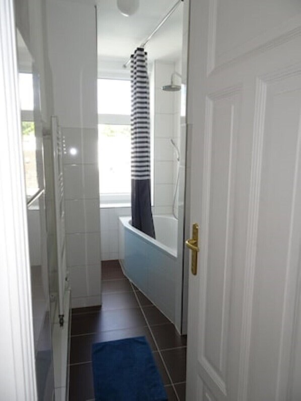 Un baño estrecho con paredes de azulejos blancos, un tapete de baño azul, una bañera blanca y una zona de ducha.