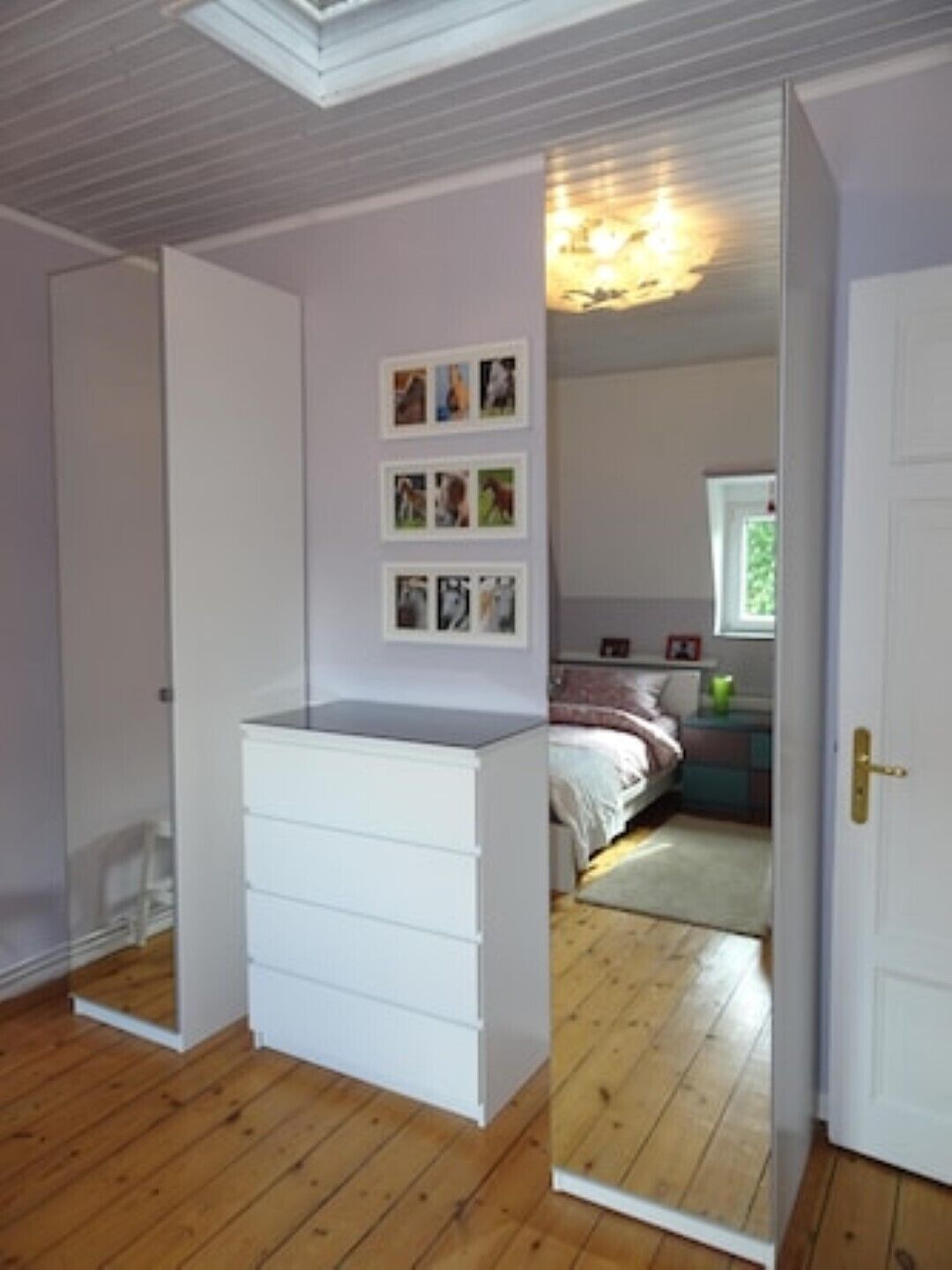 A bedroom with a wooden floor, a white dresser, a bed with a pink and white bedspread, and a window with a view of greenery.