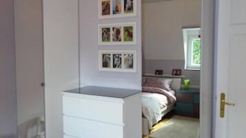 A bedroom with a wooden floor, a white dresser, a bed with a pink and white bedspread, and a window with a view of greenery.