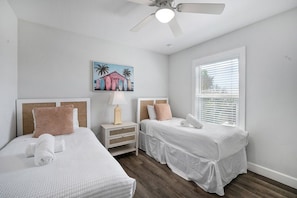 A hotel room with two beds, a ceiling fan, a painting, a window with blinds, and a nightstand with a lamp.