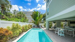 Piscina rectangular con una terraza blanca, rodeada de exuberante vegetación y una valla blanca.