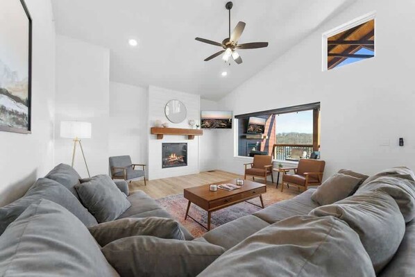A modern living room with a fireplace, a ceiling fan, and a large window offering a view. - Lake View|Pools·Hot Tub·Sauna|Game&Theater Rm·18 (Indian Point)