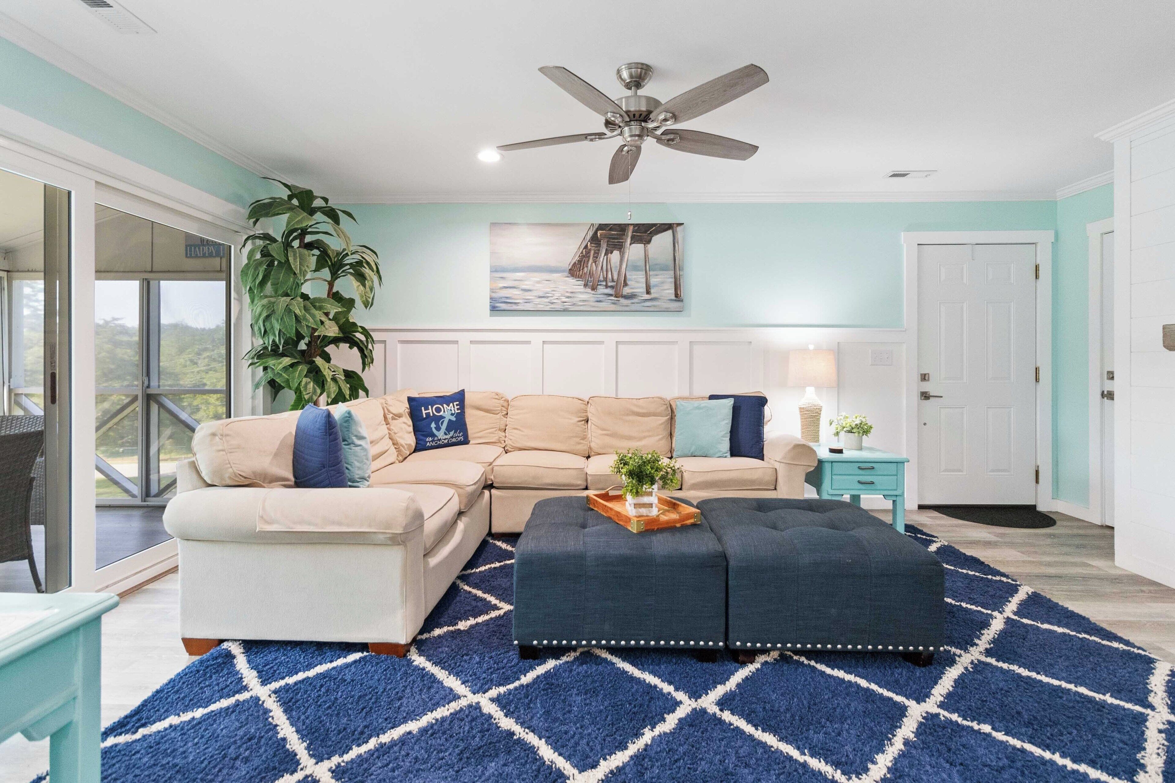 A living room with a sectional sofa, a dark ottoman, a blue rug, a ceiling fan, and a large plant.