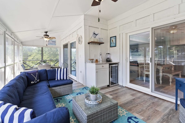 A sunroom with a large window, a ceiling fan, a kitchen area, and a dining table with chairs.