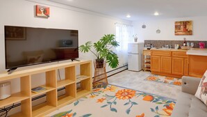 Een woonkamer met een flatscreen televisie, houten planken, een bank en een keukenhoek met houten kasten en een koelkast.