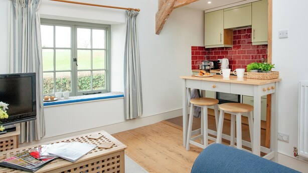 A room with a TV, a window with curtains, a kitchen area with stools, and a wooden ceiling beam.
