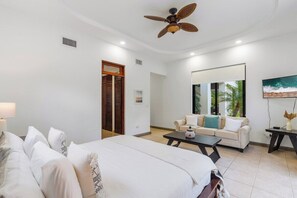 A hotel room with a bed, a sofa, a coffee table, a ceiling fan, a TV, and a painting on the wall.