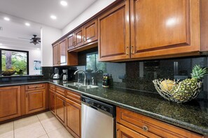 A modern kitchen with wooden cabinets, granite countertops, a dishwasher, and a window with a view of greenery.