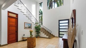 A spacious foyer with a wooden door, a staircase, a round table with a vase, and a large window with a view of palm trees.
