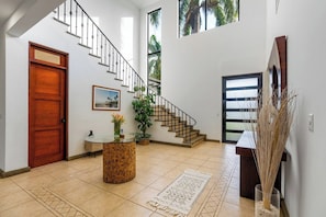 A spacious foyer with a wooden door, a staircase, a round table with a vase, and a large window with a view of palm trees.
