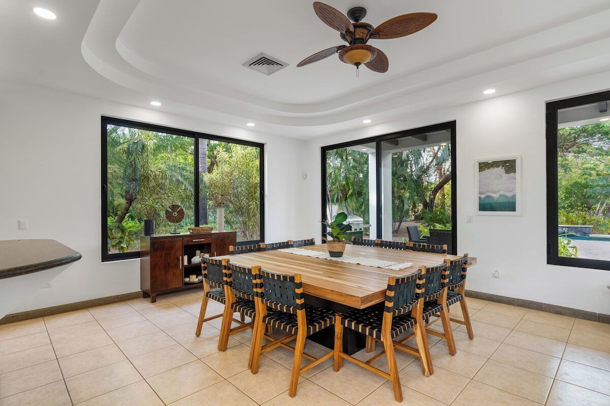 Un coin repas avec une table et des chaises en bois, de grandes fenêtres donnant sur un paysage verdoyant et un ventilateur de plafond.