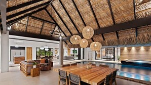 A spacious indoor area with a wooden dining table, chairs, a pool, and a thatched roof ceiling.