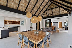 A spacious living area with a large wooden dining table, wicker chairs, and a high ceiling with a thatched roof.