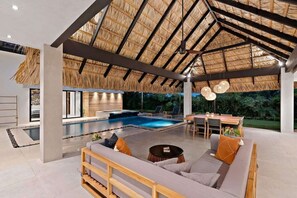 Un área de piscina interior moderna con techo de paja, piscina, asientos de descanso y zona de comedor.