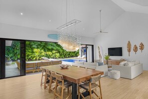 A modern living space with a dining area, a sofa set, a TV, and a view of a lush garden and pool.