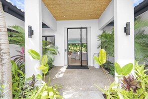 A modern building entrance with a glass door, surrounded by tropical plants, and a concrete pathway leading to a seating area. - Casa Prana (Reserva de Golf 84, Hacienda Pinilla)