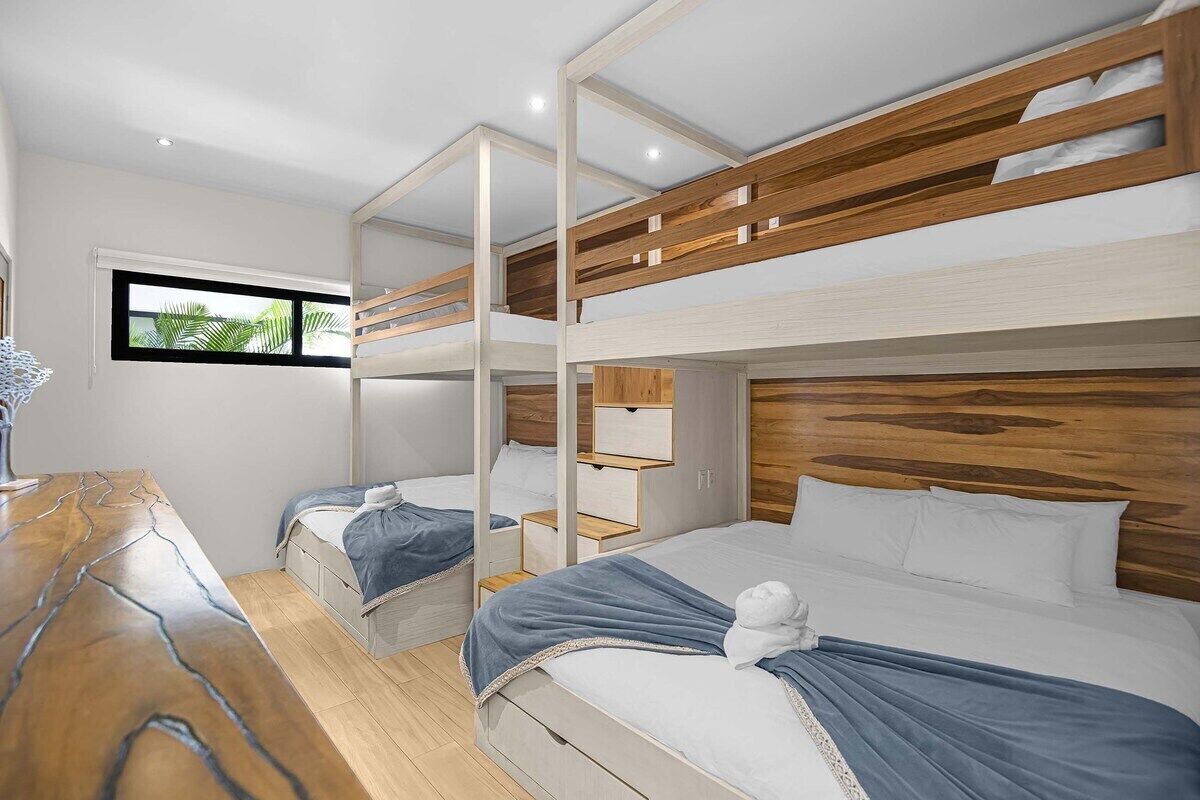 Een slaapkamer met stapelbed, een houten hoofdbord, een bureau met een vaas en een raam met uitzicht op groen.