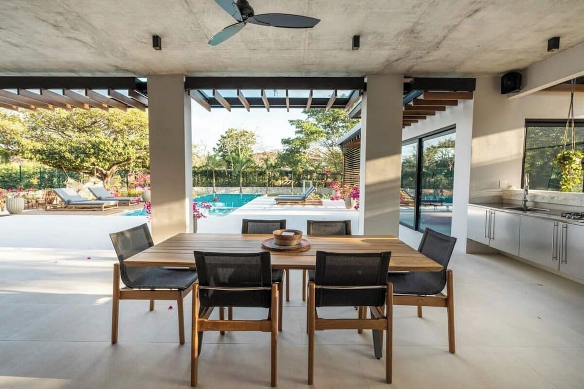 A modern outdoor dining area with a pool view, a ceiling fan, and a kitchen area with a large window.