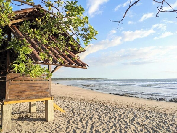 Eine Holzhütte am Strand mit einem Dach, das von Holzbalken gestützt wird, umgeben von Sand und mit Blick aufs Meer.