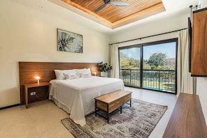 A bedroom with a bed, bedside tables, a bench, a ceiling fan, a large window, and a view of the outdoors.
