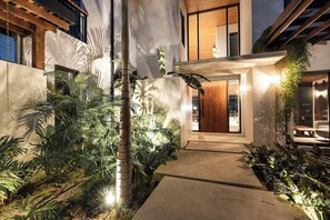 A modern house with a concrete pathway leading to a wooden door, surrounded by lush greenery. - Villa Solana (Tamarindo)