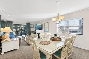 A dining area with a glass table, white chairs, and a chandelier. There is a sofa and a coffee table with a lamp in the background.