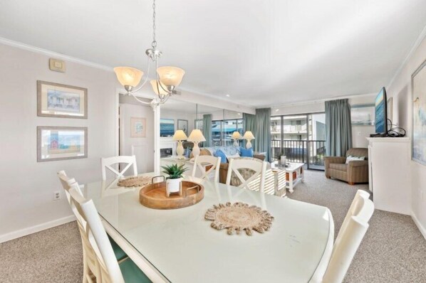 A modern living room with a glass dining table, white chairs, and a chandelier. There are framed pictures on the wall and a view of the outside through large windows.