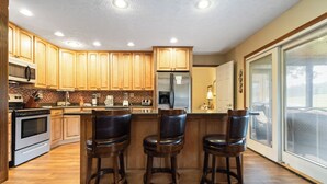 Una cocina con armarios de madera, un refrigerador de acero inoxidable, un microondas y una estufa. Hay tres taburetes de bar y una ventana con persianas.