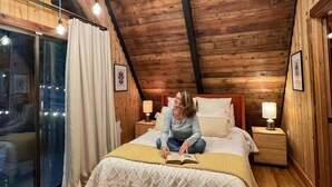 Un dormitorio acogedor con techo inclinado, paredes de madera, una cama con colcha blanca, lámparas de noche y vistas a través de un ventanal.