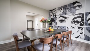 Uma sala de jantar com uma mesa comprida, seis cadeiras de madeira com almofadas cinzentas, um vaso com flores e uma parede com arte em estilo de história em quadrinhos preto e branco.