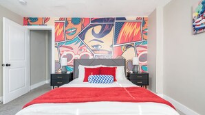 Quarto com cama, manta vermelha, almofada xadrez e parede com mural de arte pop.