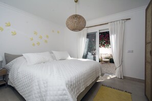 Ein Schlafzimmer mit einem großen Bett, einem Nachttisch, einer Hängelampe und Blick auf einen Balkon mit Stuhl und Pflanzen.