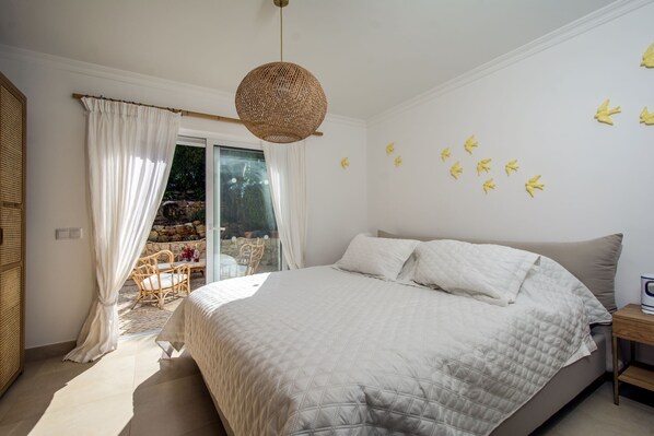 Ein Schlafzimmer mit einem großen Bett, einem Korbsessel, einem kleinen Tisch und Blick ins Freie durch eine Glastür.
