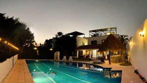 Una piscina con una estructura de techo de paja, un área de bar y una zona de estar con sillas y mesas.