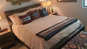 A neatly made bed with patterned pillows and a decorative throw, a bedside table with a lamp, and a window with a view of greenery. - "Cactus Cooler Casita" Private Pool and Heated Spa Included Large walk-in Shower (Queen Creek)