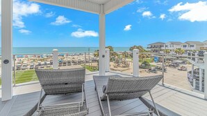 Overdekt balkon met rieten stoelen met uitzicht op een strand en parkeerplaats.