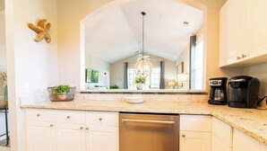 Une cuisine avec des armoires blanches, un lave-vaisselle en acier inoxydable, un comptoir en granit, un lustre et une machine à café.