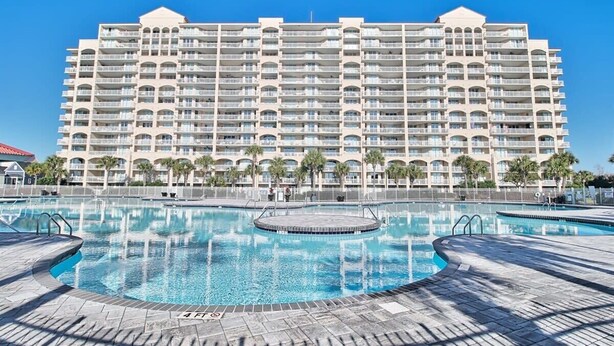 Un grand hôtel doté de nombreux balcons, d’une piscine centrale et d’un jacuzzi circulaire, le tout sous un ciel d’un bleu éclatant.