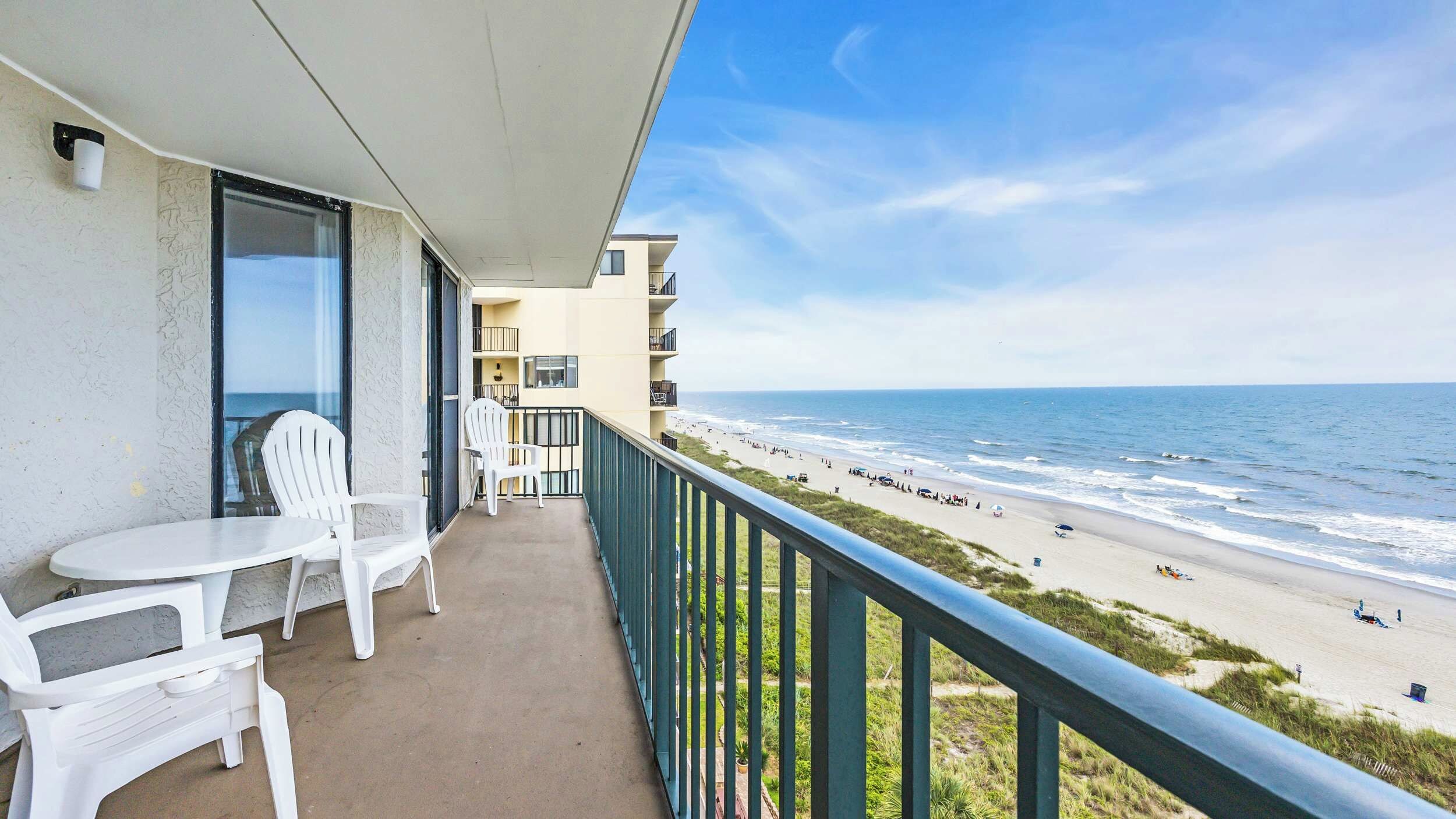 A balcony with white plastic chairs and a table, overlooking a beach with people and a clear sky.