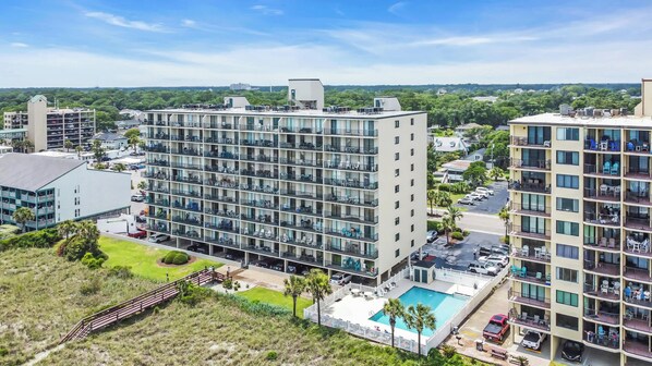 Un hotel de varios pisos con piscina en la azotea, rodeado de vegetación y otros edificios.