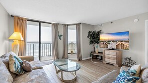 A spacious living room with a glass coffee table, a sofa, a TV, and a balcony with curtains.