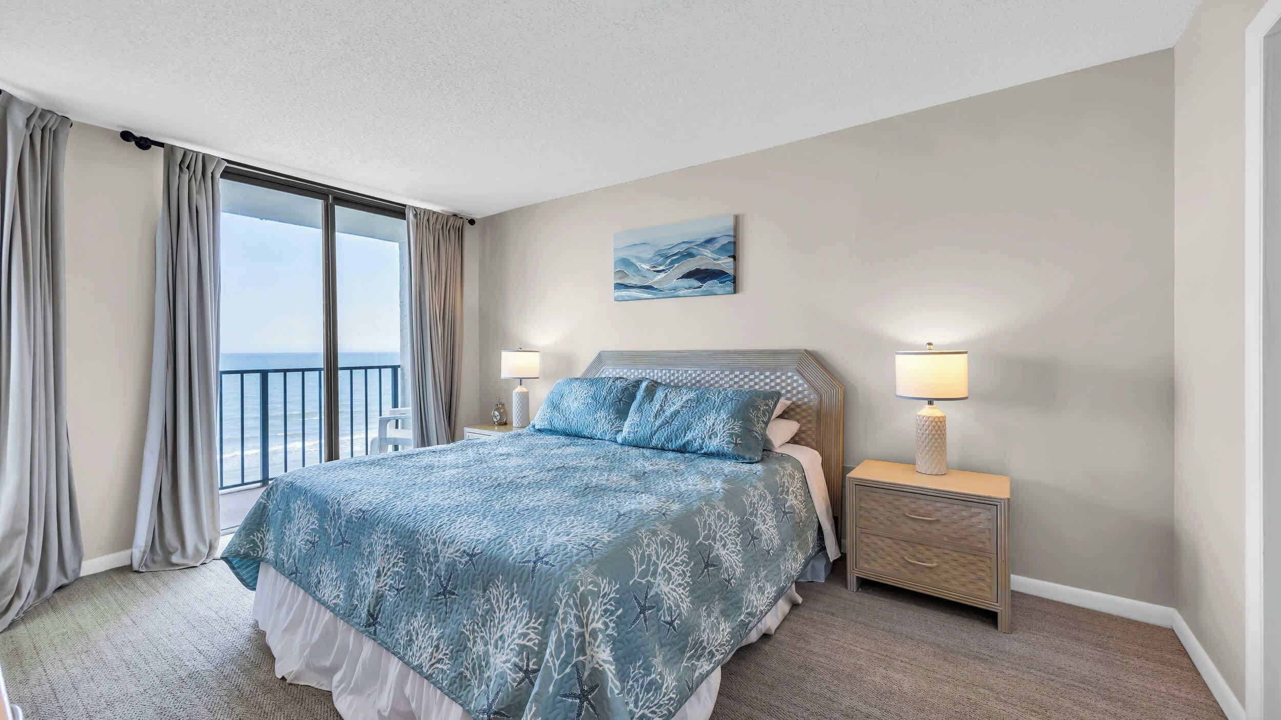 Habitación de hotel con cama, mesita de noche, lámpara, un cuadro y vista al mar a través de amplias ventanas.