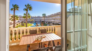 A balcony with a wooden table and chairs, overlooking a pool area with palm trees and a building complex. - Beautiful apartment in Santa Pola (Santa Pola)