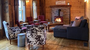 A cozy log cabin interior with wooden floors, a stone fireplace, and rustic furnishings including a sofa, armchairs, and a dining table.