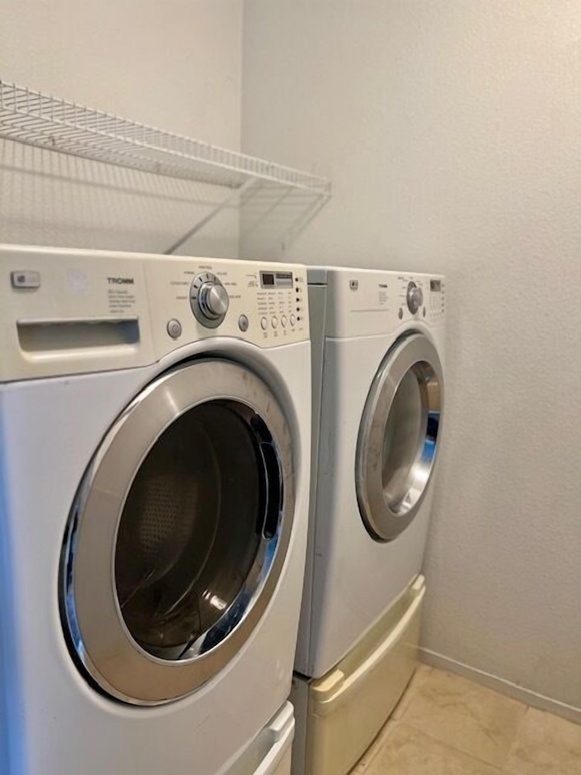 Zwei Waschmaschinen mit Frontlader in einem Waschraum mit gefliestem Boden und einem Metallregal darüber.