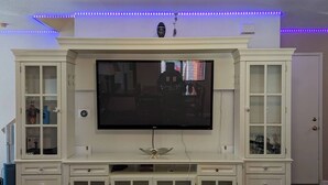 A living room with a large flat-screen TV mounted on a white entertainment center, blue LED strip lights along the ceiling, and a dark wooden floor. - By Popular Demand - Richmond (Richmond)