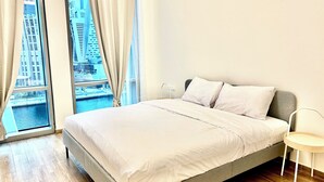 Un dormitorio moderno con una cama grande, una mesita de noche pequeña y vistas a un paisaje urbano a través de ventanales de piso a techo.