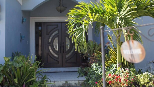 A house with a dark, ornate front door, a small porch, and various potted plants and flowers in the front yard. - 5min Walk from Love beach | Spacious Home in Nassau (Nassau)