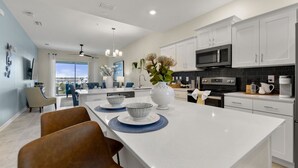 A modern kitchen with white cabinets, a central island, and a dining area with chairs. - Lake View At Storey Lake By Shine Villas #703 2 Bedroom Apts (Kissimmee)